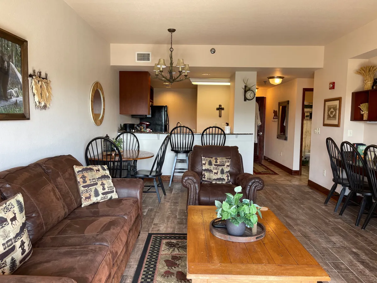 A spacious living area with a leather sofa and wooden coffee table, connected to a dining area and kitchen counter. The decor is comfortable, inviting, and ideal for socializing or relaxing. Wall art and soft lighting enhance the atmosphere.