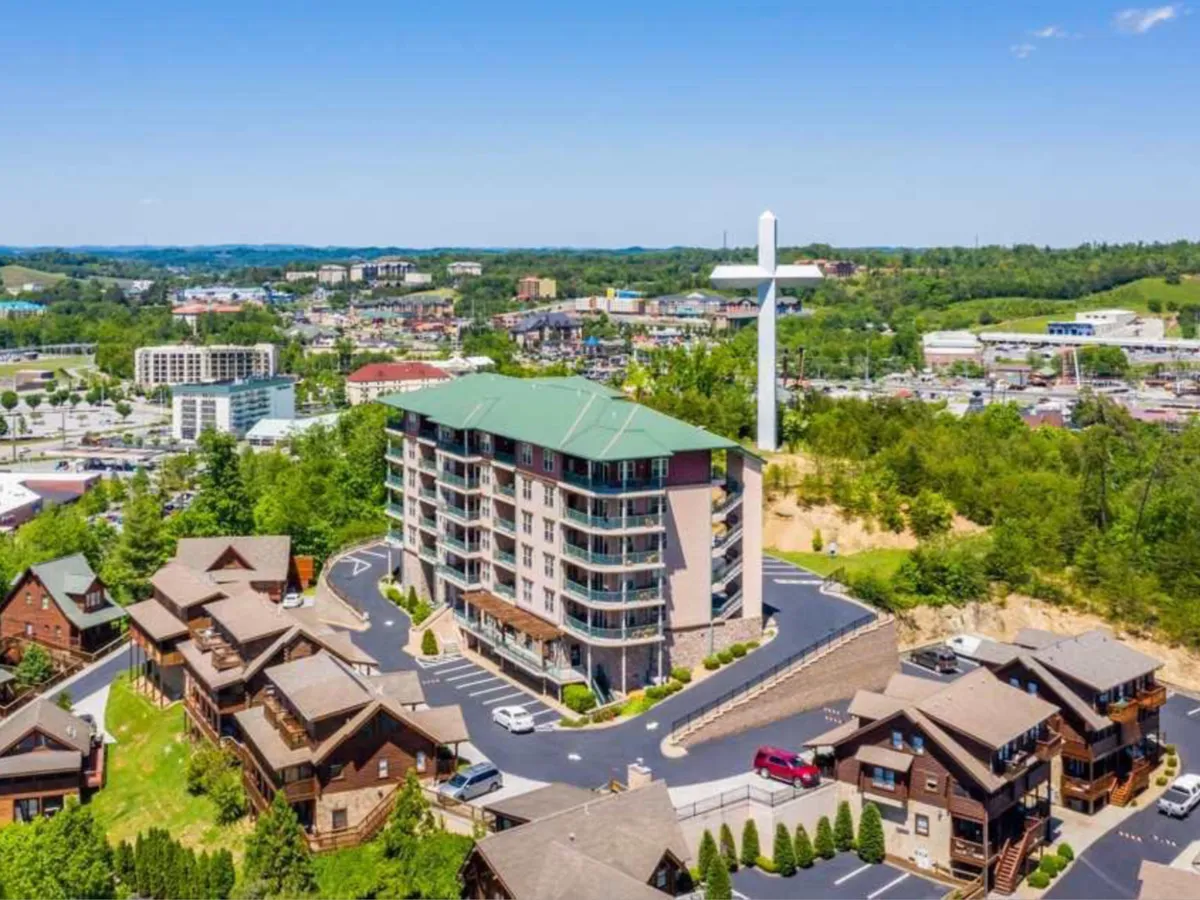 This exterior shot features a modern multi-story building surrounded by charming cabins, set against the peaceful backdrop of a large white cross on the hill. The image highlights the inviting atmosphere of the property and hints at the family-friendly community vibe, with open outdoor spaces suitable for gatherings and exploration.