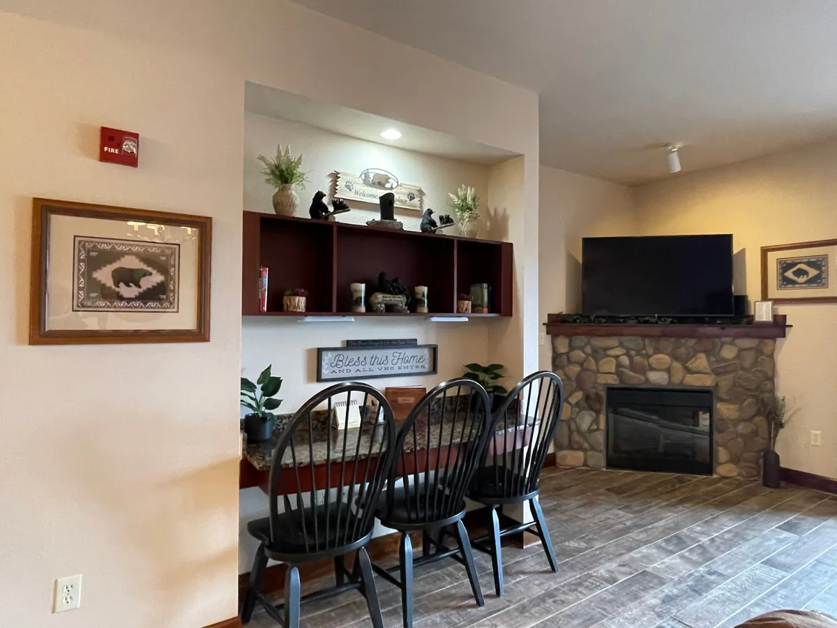 This image showcases a cozy living room featuring a prominent fireplace, a dining table with chairs, a television, and tasteful shelf decor. The space exudes warmth and lodge-style charm with wood accents and an inviting, comfortable arrangement.