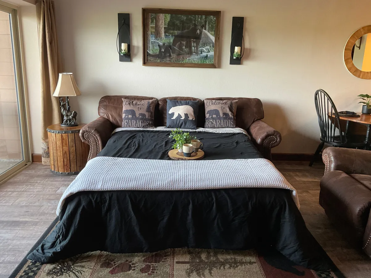 A warm, rustic living and dining area with a sofa featuring bear-themed pillows, a dining table, a lamp, and decorative nature-inspired wall art. Sunlight streams in through the window, suggesting a cozy environment for socializing and unwinding after a day of exploring local attractions.