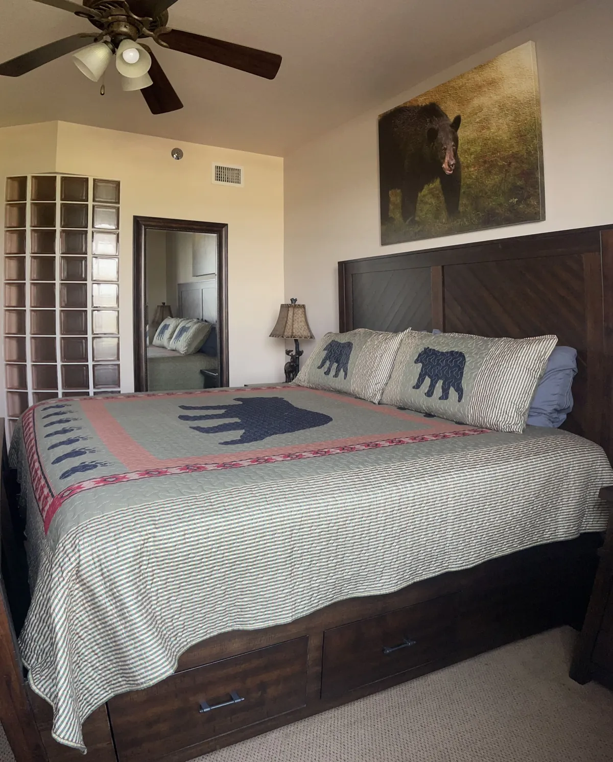 A cozy bedroom featuring a bed with a bear-themed quilt, a prominent bear portrait on the wall, and sturdy wooden furniture. The space exudes comfort and a connection to nature, reflecting a restful cabin ambiance.