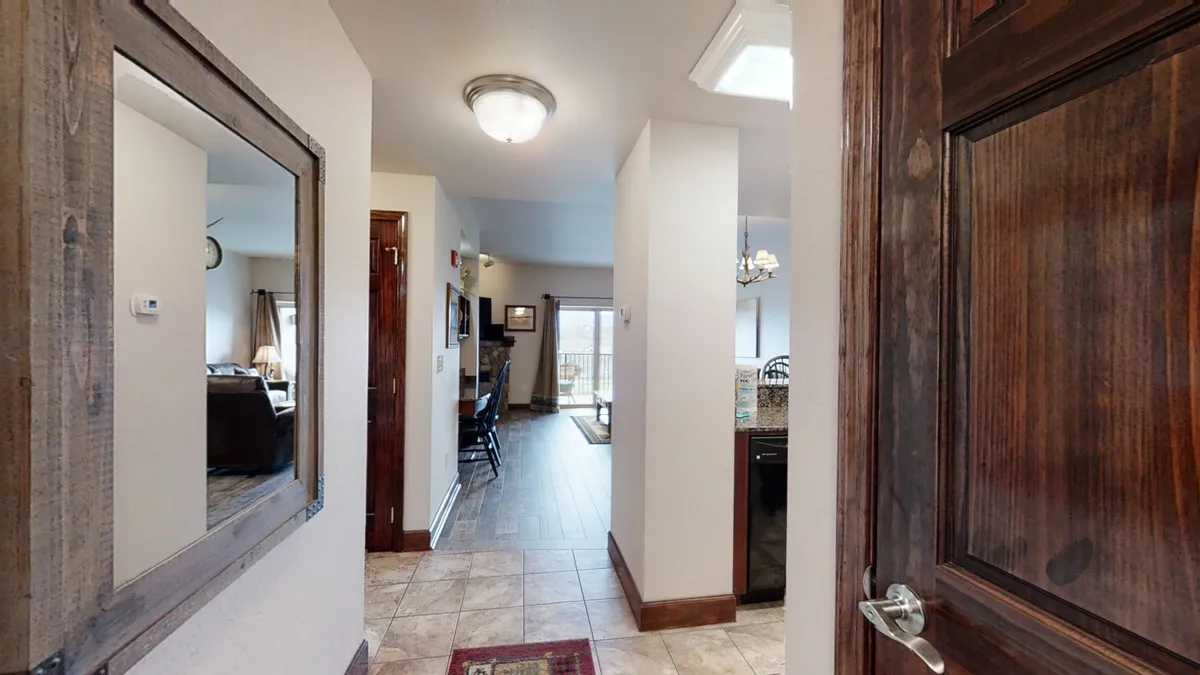 The image offers a perspective from the entryway, showing a glimpse into the open living area, including the kitchen, dining area, and living room. Visible wood details, bright natural light, and welcoming decor set a warm first impression.