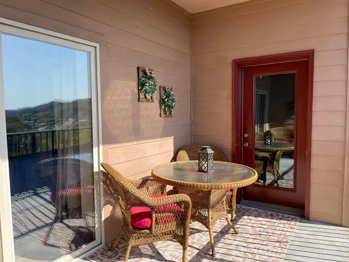 A cozy outdoor patio featuring wicker furniture, a round glass table, decorative wreaths, and a lantern, set against a backdrop of lush greenery. This inviting setup suggests a perfect spot for morning coffee or sunset drinks, connecting guests to the outdoors in comfort.