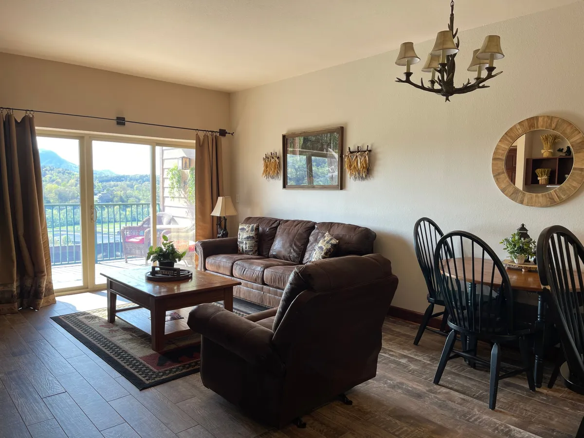 A cozy dining area is shown with a table for four, elegant chandelier, and a comfortable living space nearby. The balcony view adds natural light, creating a warm, inviting atmosphere ideal for dining or sharing stories.