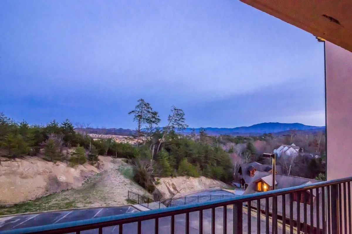 A stunning view image capturing distant mountains, a tree-lined foreground, and a gentle pathway leading down from a balcony. The scene is peaceful, ideal for morning coffee, sunset watching, or unwinding with friends after a day of adventure.