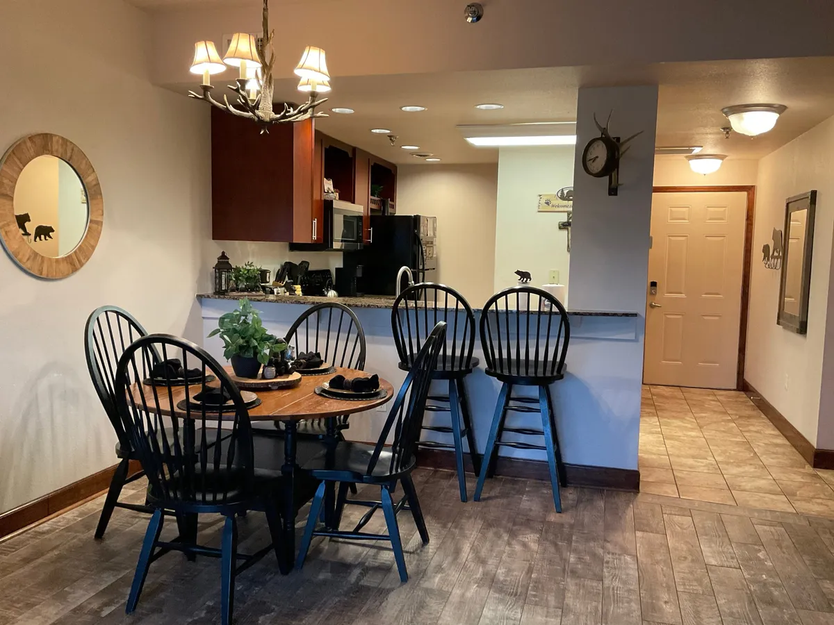 The photo focuses on a round dining table set for four, with adjacent bar stools at the kitchen counter. Appliances and a decorative mirror reflect a cozy, welcoming dining space ideal for casual meals or games.