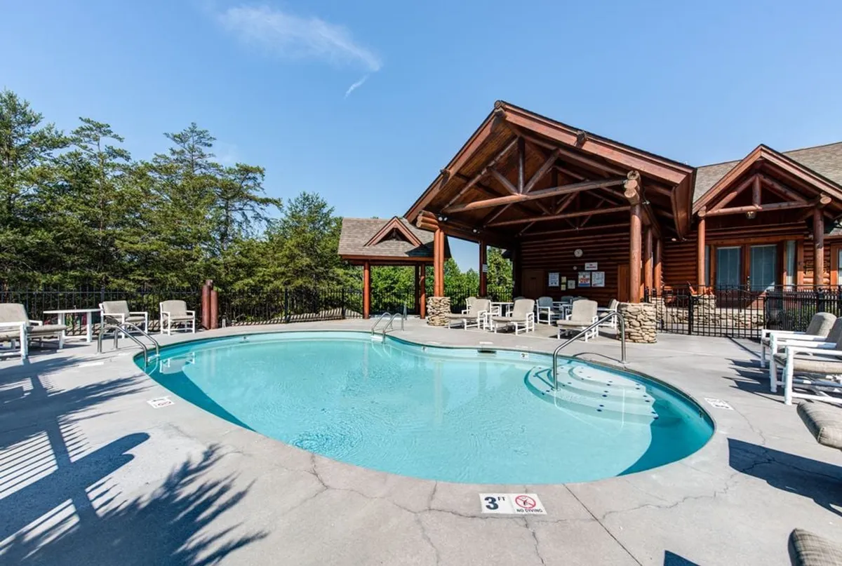 An exterior view of the resort's swimming pool area with sparkling water, lounge chairs, and a wooden structure nearby. The pool area is bathed in sunlight, evoking a relaxed, social atmosphere ideal for sunbathing, swimming, or gathering with friends.